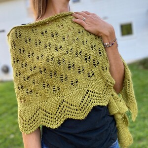 May include: A knitted shawl in a muted olive green color. The shawl features a detailed openwork pattern with a scalloped edge. The garment is draped over the shoulders, showcasing its texture and design. The shawl is a cozy accessory.