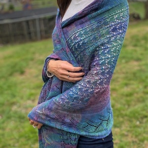 May include: A knitted shawl in shades of blue, green, and purple. The shawl has a delicate, openwork design and is draped over the shoulders. The colors blend together, creating a gradient effect. The shawl is worn over a white top and dark blue jeans.