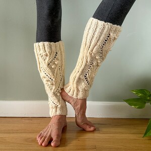 May include: Cream-colored knitted leg warmers with a textured pattern and bobble details. The leg warmers are worn on the legs. The leg warmers are made of a soft, warm material. The leg warmers are a cozy accessory for cooler weather.