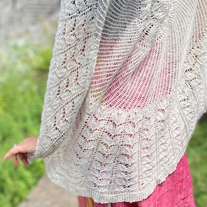 May include: A light beige, open-weave shawl with intricate leaf and lattice patterns. The shawl is draped over the shoulders, showcasing the delicate knit design. The edges feature a scalloped border, adding a touch of elegance to this handcrafted accessory.