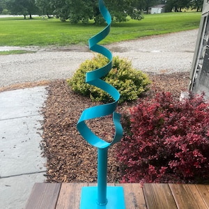May include: A teal blue metal sculpture of a spiral shape on a square base. The sculpture is outdoors on a wooden deck.
