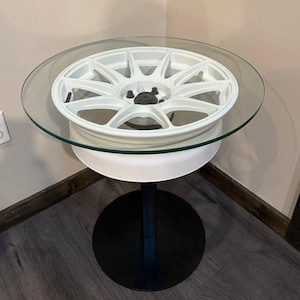 May include: A circular table featuring a white automotive wheel rim as the base, topped with a clear round glass surface. The table is supported by a vertical black metal post connected to a solid, circular black metal base plate on the floor.