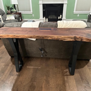 May include: A live edge walnut wood console table with black metal legs. The table has a natural wood finish and a live edge design. The table is approximately 5 feet long and 18 inches wide.