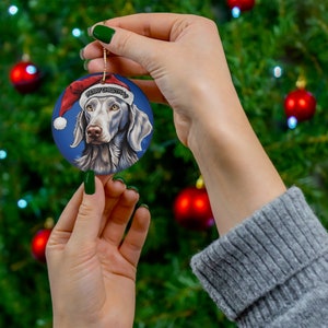 Op de afbeelding: Een ronde kerstbal met een Weimaraner die een kerstmanmuts draagt. De kerstbal heeft een blauwe achtergrond met de tekst "Merry Christmas" erop.