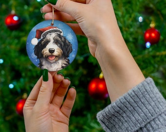 Op de afbeelding: Een ronde kerstversiering met een hond met een kerstmuts, met de tekst "Merry Christmas" op de muts. De ornament heeft een blauwe achtergrond met sneeuwvlokken en een gouden touwtje om op te hangen.