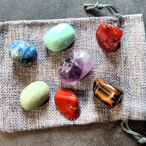 Peut inclure: Une pochette en tissu gris contenant sept pierres roulées : verte, bleue, violette, rouge et marron. Les pierres sont disposées en demi-cercle.