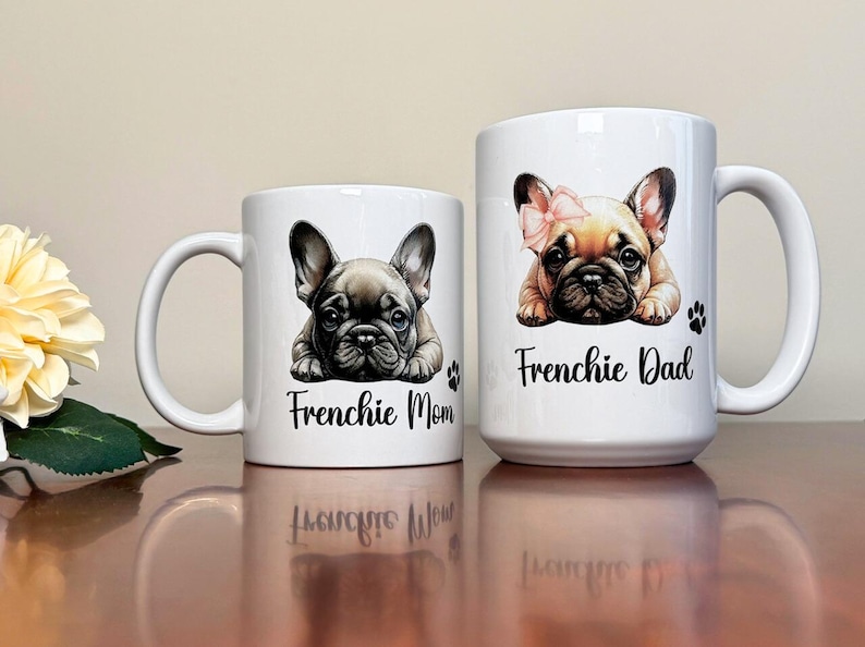 Left mug shows a gray blue French Bulldog while on the right mug shows a Fawn French Bulldog with with pink and black color ears. Left mug says Frenchie Mom and Right mug says Frenchie Dad with a paw print.