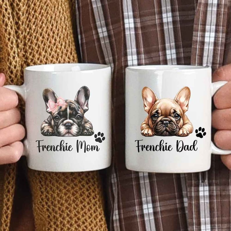 The left mug shows a Brindle French Bulldog with a bow with a text says Frenchie Mom with a paw print next to the dog. The right mug shows a Fawn French Bulldog without a bow with a text says Frenchie Dad and a paw print next to the dog.