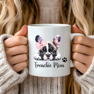 A 11oz white mug watercolor pied French Bulldog mug with a paw and a text says Frenchie Mom. The French Bulldog is lying on the floor with a puppy eye look.