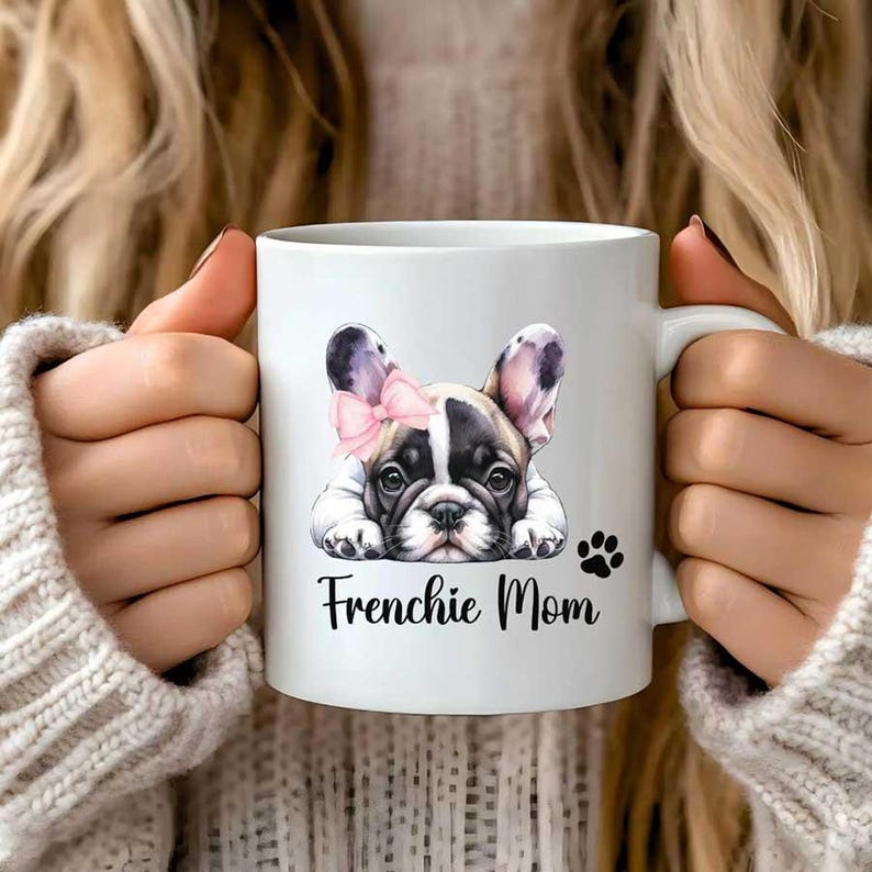 A 11oz white mug watercolor pied French Bulldog mug with a paw and a text says Frenchie Mom. The French Bulldog is lying on the floor with a puppy eye look.