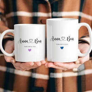 Puede incluir: Dos tazas de cerámica blanca con letras negras. Las tazas están personalizadas con los nombres "Anna" y "Ben" y la fecha "14 de febrero de 2025". Una taza tiene un corazón rosa y la otra un corazón azul.