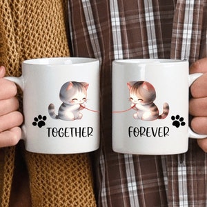 May include: Two white ceramic mugs featuring a cute cartoon cat design. The mug on the left reads "Together" and the mug on the right reads "Forever".