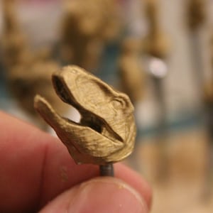 May include: A small, gold-colored dinosaur head with an open mouth, held by a person. The head is detailed with textured surfaces and appears to be a miniature sculpture or figurine. The background is blurred.