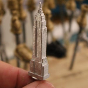 May include: A detailed silver miniature of the Empire State Building. The model is held in a hand, with other blurred objects in the background. The building has intricate architectural details, including windows and a spire.