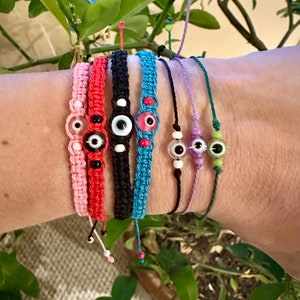 May include: Seven colorful string bracelets with evil eye charms. The bracelets are made of different colors of string, including pink, red, black, blue, purple, and green. Each bracelet has a small evil eye charm with a blue eye and a white pupil.