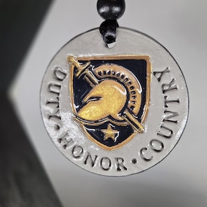 May include: A round, silver-colored pendant with the words "DUTY HONOR COUNTRY" encircling a gold and black emblem. The emblem features a helmet and crossed swords. The pendant is suspended by a black cord with gold and black beads.