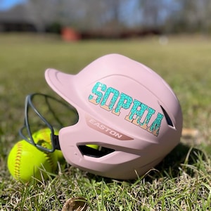 May include: A pink softball helmet with the name "SOPHIA" in turquoise and tan lettering. The helmet has a face guard and sits next to a yellow softball on a grassy surface. The brand "EASTON" is visible on the helmet.