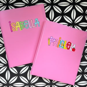 May include: Two pink folders with the names "Isabella" and "Paisley" in colorful, playful lettering. The "Isabella" folder has rainbow-colored letters. The "Paisley" folder has a pencil, apple, and other school-themed illustrations.