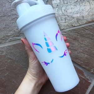 May include: White shaker bottle with a unicorn design featuring a pink and blue iridescent horn and eyelashes.