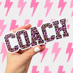 May include: A white sticker with the word "COACH" in a pink leopard print design with gold accents. The sticker is held in a hand against a background of pink lightning bolt shapes.