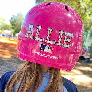 May include: Pink baseball helmet with the name "ALLIE" in white letters with red stitching on the front. The helmet has a Rawlings logo on the side.