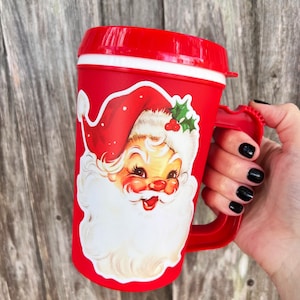 May include: Red and white Christmas mug with a Santa Claus illustration. The mug has a handle and a white rim.