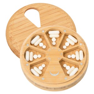 Peut inclure: Un pilulier rond en bambou avec un couvercle pivotant doté d'une poignée découpée. L'intérieur est divisé en huit compartiments en forme de coin contenant divers médicaments blancs sous forme de gélules et de comprimés. Une lune est gravée.