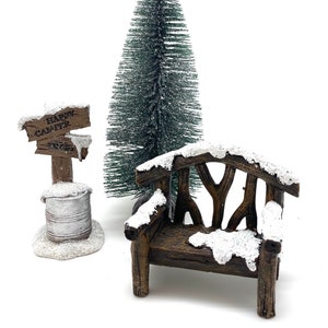 May include: A miniature winter scene with a snow-covered wooden bench, a small evergreen tree, and a sign that reads "Happy Camper" with an arrow pointing to the right.