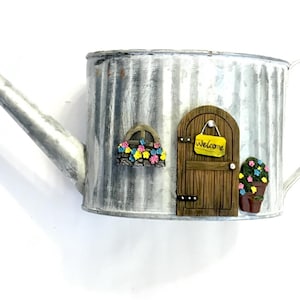 May include: A galvanized metal watering can with a decorative fairy garden theme. Features a wooden door with a "Welcome" sign, a window box with colorful flowers, and a potted plant. The watering can has a spout and handle.