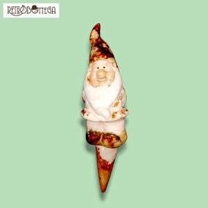 Può includere: Un gnomo da giardino in ceramica con la barba bianca e il cappello marrone. Lo gnomo è in piedi su un paletto marrone e bianco.