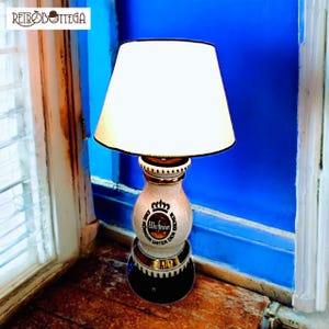 May include: A white lampshade with a black trim sits atop a ceramic lamp base. The base is decorated with a gold band and a circular design featuring the text "Warsteiner", "König", "Unter", and "Den", in a gold and black font.