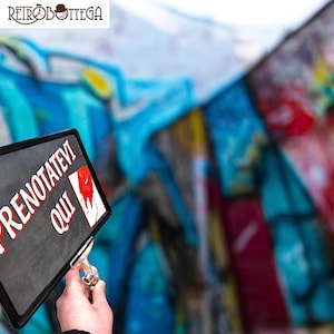 Könnte beinhalten: Ein schwarzes Schild mit weißem Text, der "Prenotatevi Qui" lautet, mit einer roten Hand, die auf den Text zeigt. Das Schild wird vor einer bunten, mit Graffiti besprühten Wand gehalten.