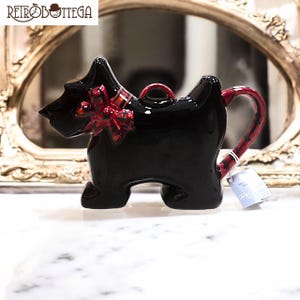 Könnte beinhalten: Schwarze Keramik-Teekanne in Form eines Scottish Terrier-Hundes, mit roter Schleife und Griff. Die Teekanne hat eine glänzende Oberfläche und ein angebrachtes Etikett. Die Teekanne wird auf einer weißen Oberfläche präsentiert. Ca. 20 cm lang.