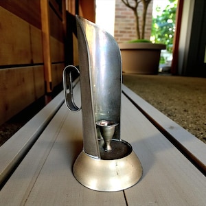 Può includere: Un portacandele vintage color argento con manico. Il portacandele ha un retro curvo e una piccola coppa per una candela. La base è arrotondata. Il portacandele è su una superficie di legno grigio.