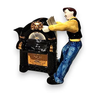 Könnte beinhalten: Eine Keramikfigur eines Mannes in einer gelben und schwarzen Jacke und blauen Jeans, der sich an einer schwarzen Jukebox mit goldenen Akzenten abstützt. Die Jukebox hat eine goldene Lautsprecherabdeckung und eine kleine Kette. Der Mann hält ein Papier.
