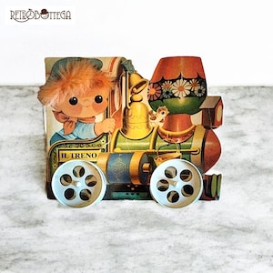 May include: A vintage cardboard toy train with a colourful design. The train has a blue and gold engine, a red caboose, and a white figure of a child with orange hair. The train has the words "IL TRENO" printed on the side.