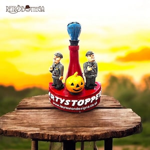 Può includere: Un tappo per bottiglia rosso e bianco con una zucca e due figure in uniforme militare. Il tappo per bottiglia dice "Partystopper" e "www.laurestonedesigns.co.uk".
