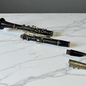 Puede incluir: Dos clarinetes negros con llaves y detalles plateados se exhiben sobre una superficie de mármol blanco. Un clarinete está completamente ensamblado, mientras que el otro está parcialmente desmontado, con su boquilla y barrilete separados.