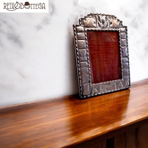 Puede incluir: Un marco de fotos de plata con un diseño ornamentado. El marco tiene un borde elevado con un patrón texturizado y una  paspartú de terciopelo rojo.