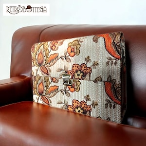 Może przedstawiać: Wzorzysta kopertówka w stylu vintage z ozdobnym złotym zamkiem. Kopertówka leży na brązowej skórzanej sofie.