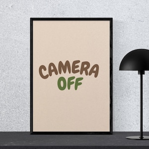May include: A framed print with the words "CAMERA OFF" in brown and green lettering on a beige background. A black desk lamp with a rounded shade is to the right of the framed print.