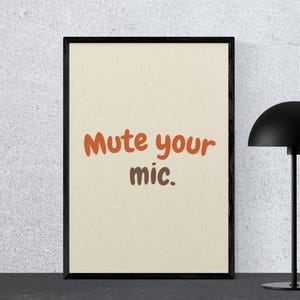 以下が含まれることがあります： オレンジと茶色で「Mute your mic.」と書かれた額入りのプリント。プリントは黒いフレームに入っており、暗い表面に置かれています。丸みを帯びたシェードの黒いデスクランプがプリントの右側にあります。