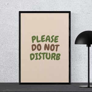 May include: A framed print with the words "PLEASE DO NOT DISTURB" in green and brown letters on a beige background. A black lamp with a dome-shaped shade is to the right of the frame.