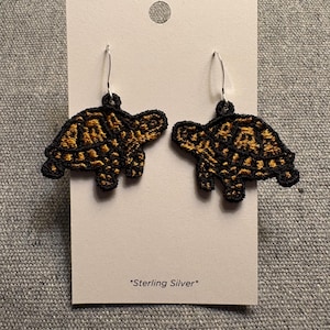 以下が含まれることがあります： 黒と金茶色の模様の刺繍されたカメのイヤリング。イヤリングはシルバーのフックに吊り下げられ、白いカードに表示されています。カードには「Sterling Silver」と書かれています。