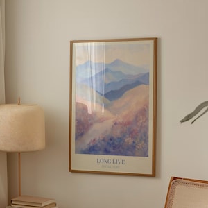 以下が含まれることがあります： 薄い青、紫、ピンクの色合いの水彩画で描かれた山脈の額装されたプリント。プリントには、下部に「LONG LIVE」と「STAY. NOW.」の文字が印刷されています。