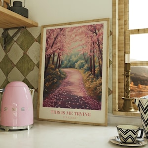 Könnte beinhalten: Ein rosa elektrischer Wasserkocher mit silbernem Griff und Sockel steht auf einer weißen Arbeitsplatte neben einem gelben Toaster und einem gerahmten Druck eines rosa-grünen Waldweges mit dem Text "This is me trying folklore".