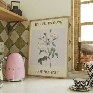 May include: A kitchen scene featuring a light pink electric kettle and a yellow toaster on a white countertop. A framed botanical print with the text "IT'S HELL ON EARTH TO BE HEAVENLY" hangs on the wall. A black and white patterned cup and saucer are also visible.