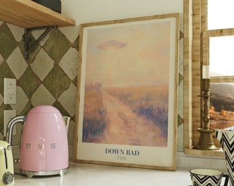 May include: A pink electric kettle with a silver handle and spout sits on a white countertop next to a yellow toaster. A framed print with a pastel landscape and the text "DOWN BAD TTPD" hangs on the wall behind the appliances.