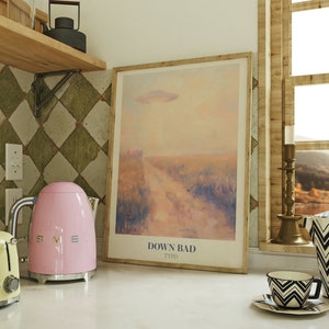 May include: A pink electric kettle with a silver handle and spout sits on a white countertop next to a yellow toaster. A framed print with a pastel landscape and the text "DOWN BAD TTPD" hangs on the wall behind the appliances.