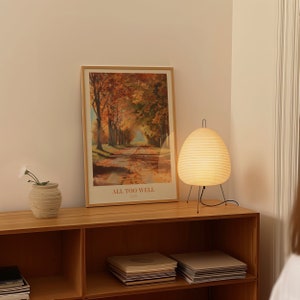 May include: A wooden shelf unit with a framed poster of an autumnal scene with the text "ALL TOO WELL (10 Minute Version)" and a white, round, hanging lamp with a black cord and stand.